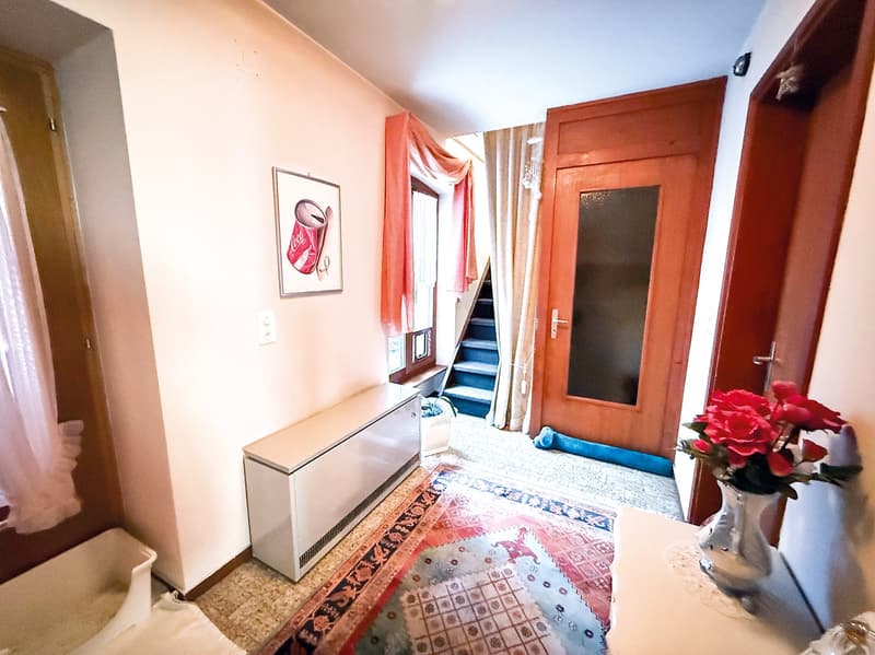 Hallway with wooden doors, a framed artwork on the wall, a cabinet, and a patterned rug on the floor. The hallway leads to a staircase and has a curtain covering a window or door.