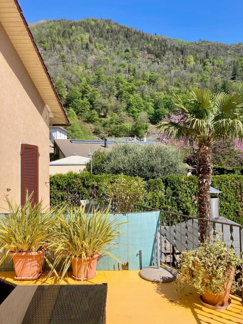 balcony, metal railing, plants, small table, house and mountains in the background