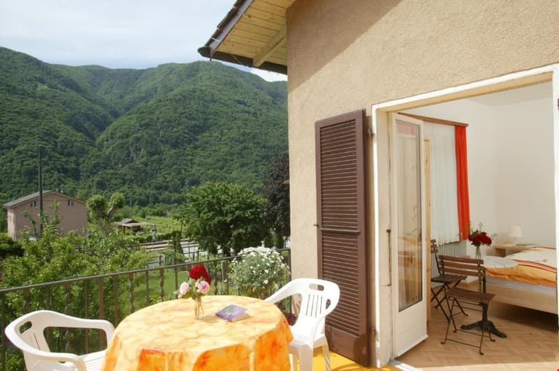 Balcony with a view of the mountains, a wooden table and white chairs, a single bed, and a flower vase