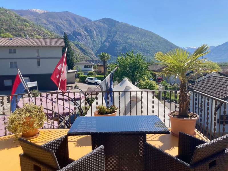 Terrace with table and chairs, fencing, potted plants, flags, view of mountains and houses.