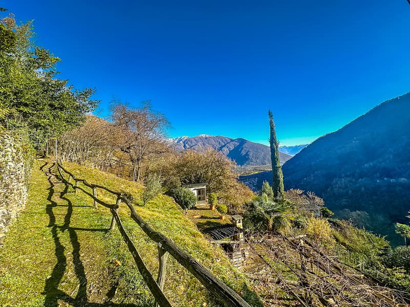 a hillside with a wooden fence, grassy area, small house, trees, mountains in the distance