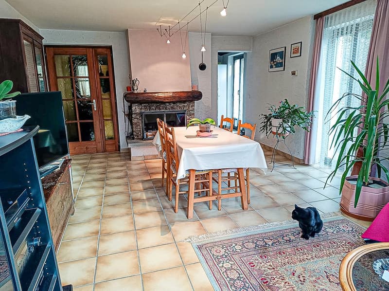 The image shows a cozy and rustic-style living and dining area. The room features a stone fireplace, wooden furniture including a dining table with chairs, and various decorative elements like plants and artwork. The floor is tiled, and there is a large w