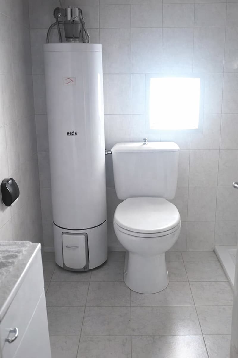 Bathroom with white tiles, white toilet, white water heater, white cabinet, and a window.