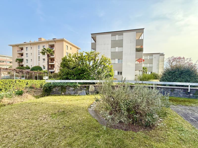 A garden with a grassy lawn, a small path, and a stone wall with a metal railing. There are trees and bushes in the garden, and a building with balconies and windows in the background.