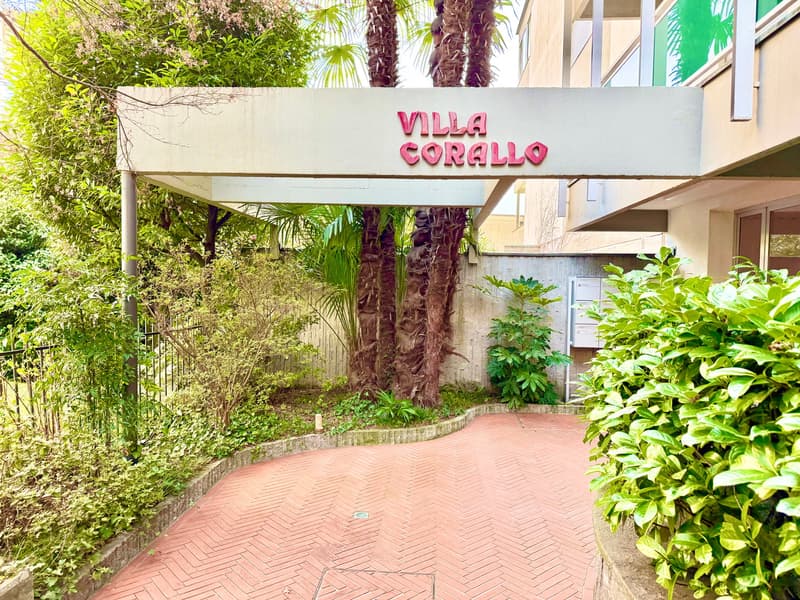 Entrance to a building named Villa Corallo, brick pathway, greenery, trees, and plants