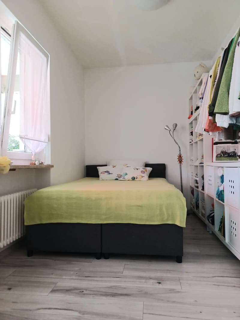 bedroom with bed, pillows, green bedspread, white walls, wooden floor, window with white curtain, white radiator, white shelves with clothes