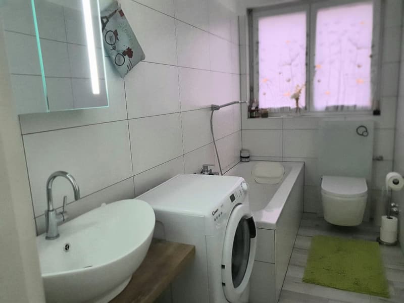 white tiled walls, white sink, white washing machine, bathtub, toilet, window, green rug