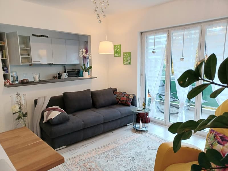 modern living room with gray sofa, wooden table, white rug, kitchen area, balcony with sliding glass doors, white walls, chandelier