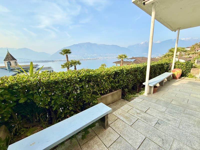 Outdoor area with a view of the mountains and lake, two white benches, and a hedge.