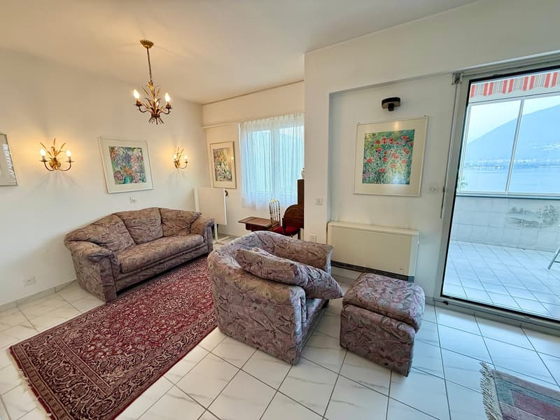 white walls, white tile floor, chandelier, two couches, two armchairs, ottoman, rug, paintings, sliding glass door to balcony, view of lake and mountains