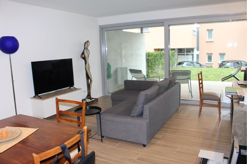 modern living room with gray sofa, wooden table, TV, glass doors leading to balcony, wooden chairs, and a view of the outside