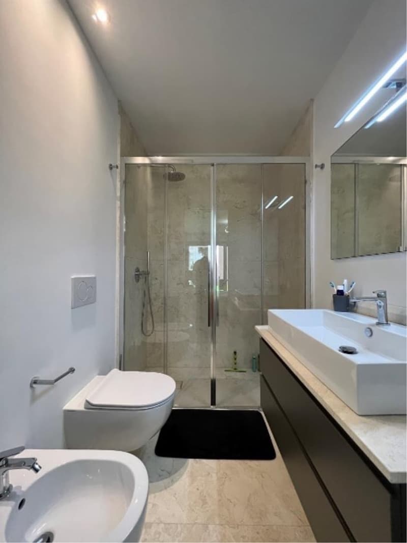 modern bathroom with white walls, marble flooring, glass shower enclosure, white sink, white toilet, mirror, black mat