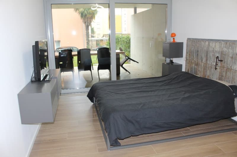 Bedroom with bed, TV, sliding glass doors, wooden floor, nightstands, and a view of a patio with chairs and a table.