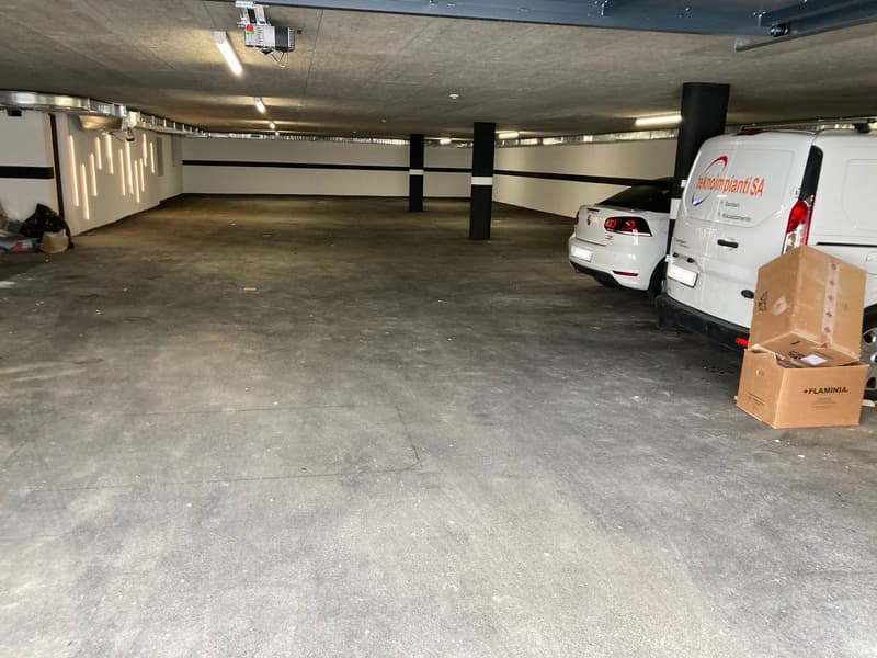Empty parking lot with two cars parked, concrete floor, white walls, ceiling lights, pillars, boxes on the right side