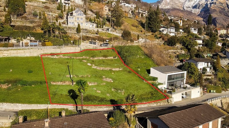 Aerial view of a plot of land with a house on the right side, surrounded by other houses and greenery, with a mountain in the background.