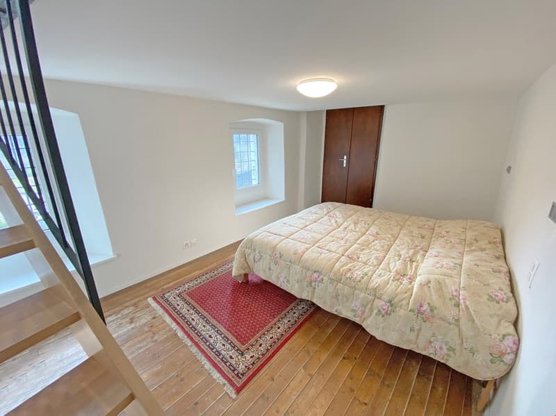 bedroom with bed, rug, wooden floor, white walls, wooden door, small window, staircase