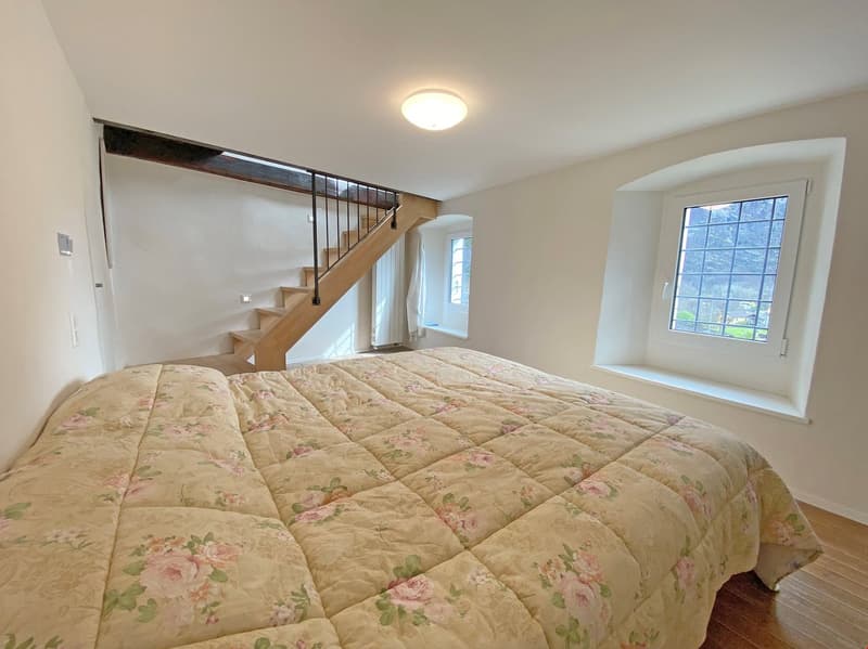 bedroom with bed, wooden floor, white walls, wooden staircase, window, ceiling light