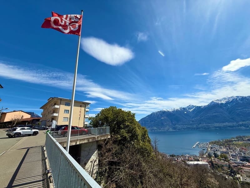 Building with a flag, overlooking a lake and mountains, with cars parked in front