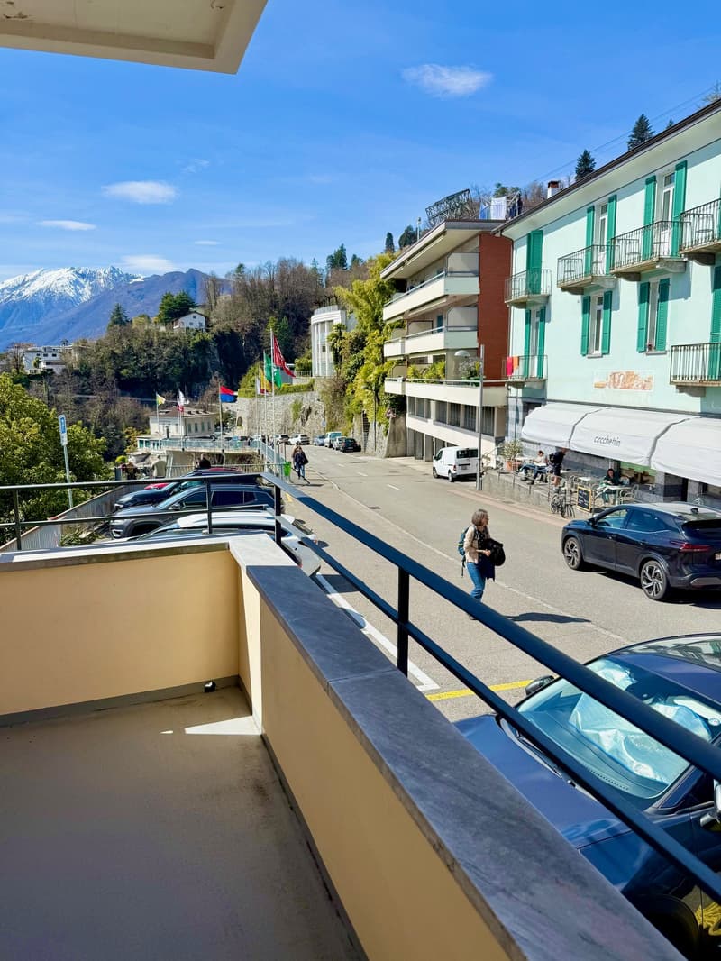 balcony with railings, cars parked on the street, people walking, mountains in the background