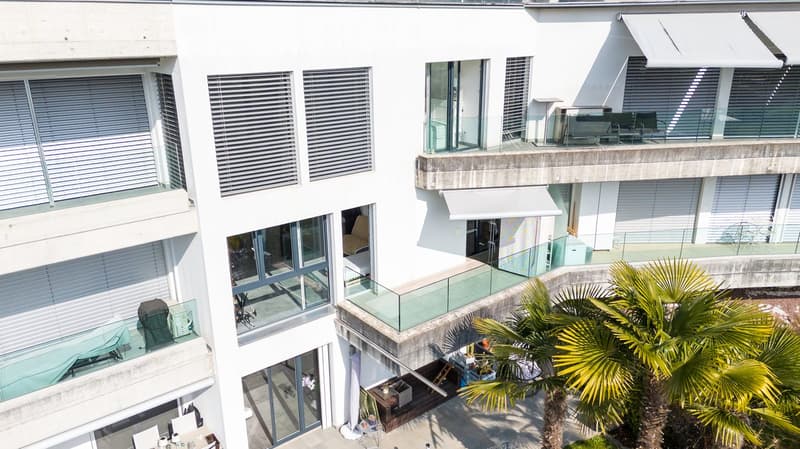 Modern white building, multiple floors, glass windows, balconies, awnings, outdoor seating area, palm trees