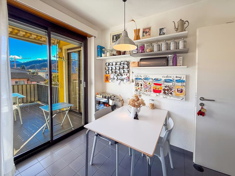 Dining area with a table and chairs, sliding glass door leading to a balcony, white walls, tiled floor, shelves with various items, a teapot, and a painting on the wall.