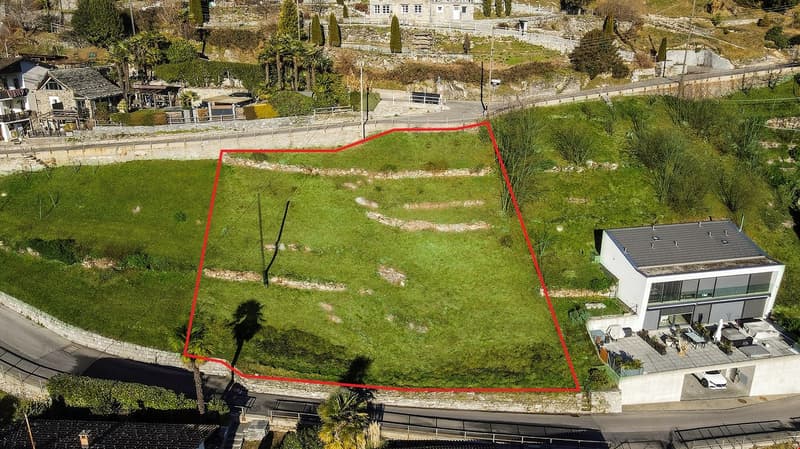 House with a garage, a car parked in front, a grassy field with a red outline, a road, and a house in the distance.