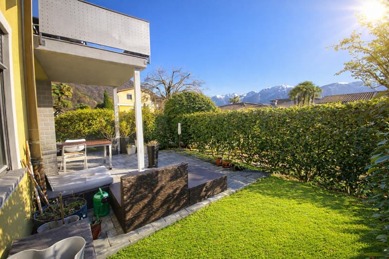 Outdoor seating area with a table and chairs, a couch, and a balcony with a railing. The garden has a lawn, hedges, and potted plants. The view includes mountains in the distance.