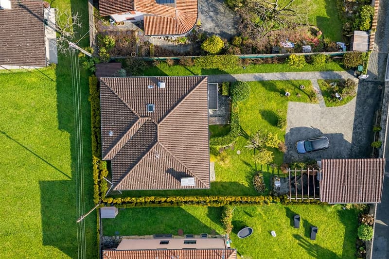 Aerial view of a house with a brown roof, surrounded by a well-maintained garden with green grass and bushes. The house is located in a suburban area with other houses and a driveway leading to the front entrance. The house has a car parked in front of it