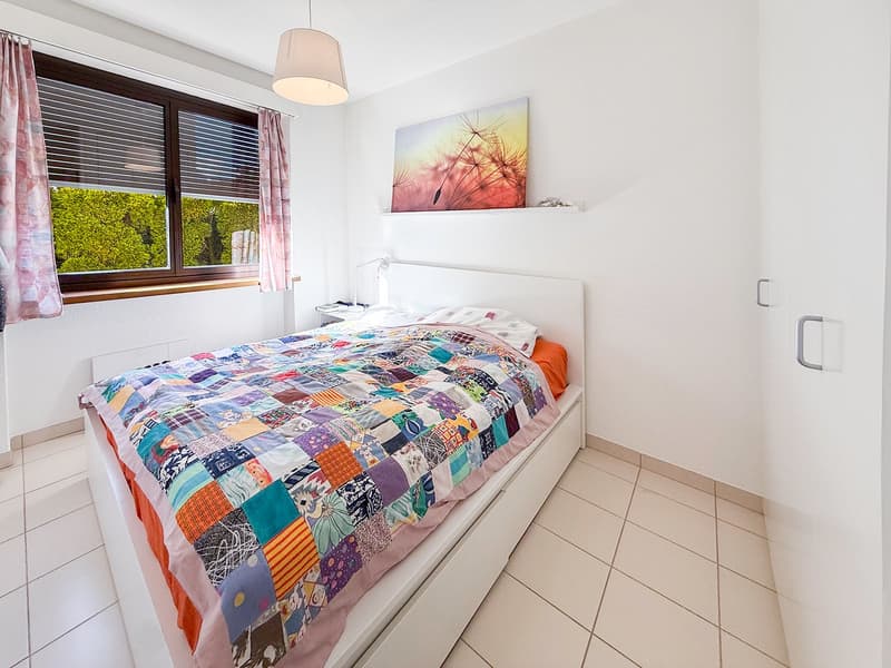 Bedroom with a bed, nightstand, lamp, and a window with blinds. Wall-mounted shelf with a picture. White walls and tiled floor.