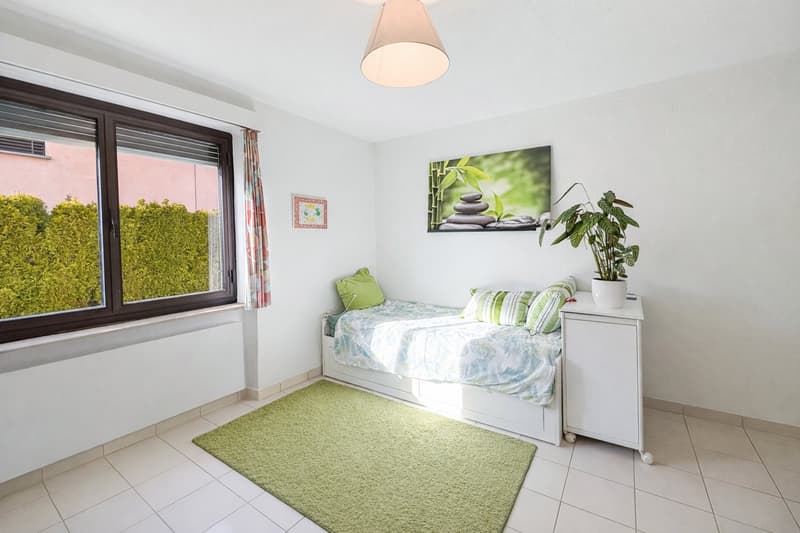 small bedroom with a single bed, white walls, green rug, white nightstand, large window, picture on the wall, plant