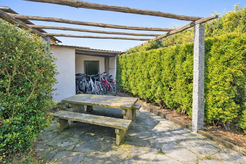 Outdoor area with a wooden pergola, a stone table, and benches. Bicycles are parked in the garage.