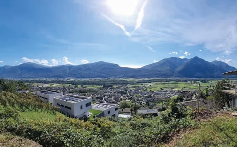 The image shows a panoramic view of a town or city nestled in a valley surrounded by mountains. The landscape features a mix of residential and commercial buildings, with green fields and trees in the foreground. The sky is clear and bright, with some clo
