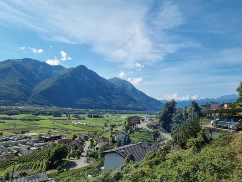 The image shows a scenic mountain landscape with rolling hills, lush green vegetation, and a small town nestled in the valley below. The sky is clear and blue, with some fluffy white clouds. The mountains in the background appear rugged and steep, creatin