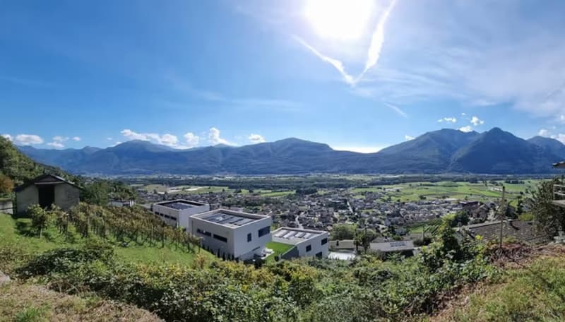 The image shows a panoramic view of a town or city surrounded by mountains. The town has a mix of residential and commercial buildings, with a modern-looking building visible in the foreground. The landscape is lush and green, with trees and vegetation co