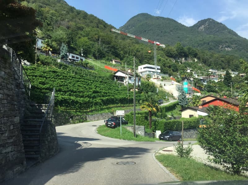 The image shows a scenic mountain landscape with a winding road, residential buildings, and lush greenery. There are several buildings with balconies or terraces visible on the hillside. The mountains in the background are covered in dense vegetation, cre