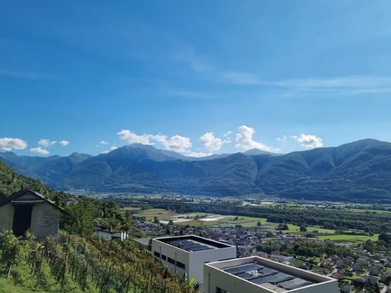 The image shows a panoramic view of a mountainous landscape with a town or village nestled in the valley below. The mountains in the background are tall and rugged, with a blue sky dotted with clouds overhead. The foreground includes some buildings or str