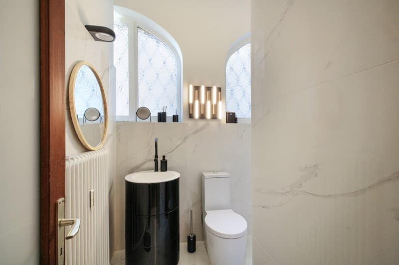 Bathroom with white marble walls, arched windows, oval mirror, modern fixtures, white radiator, modern sink, modern toilet