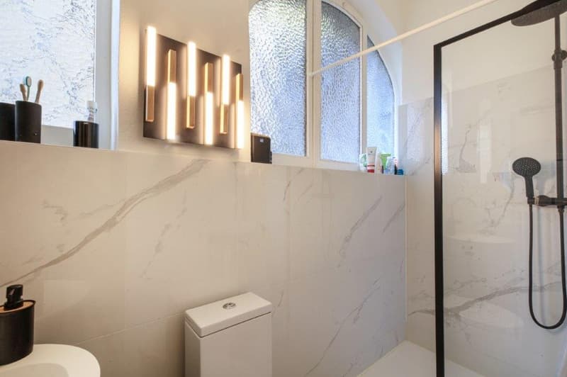 modern bathroom with white marble tiles, glass shower enclosure, white toilet, white sink, wall-mounted lights, arched windows