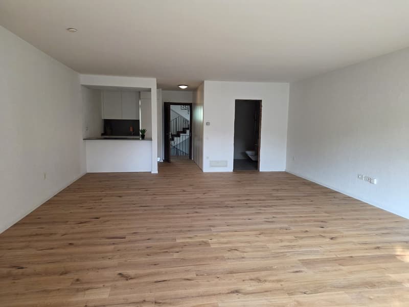 Empty room with wooden floor, white walls, kitchen area with cabinets, open door leading to staircase, another door leading to bathroom, power outlets on the wall.