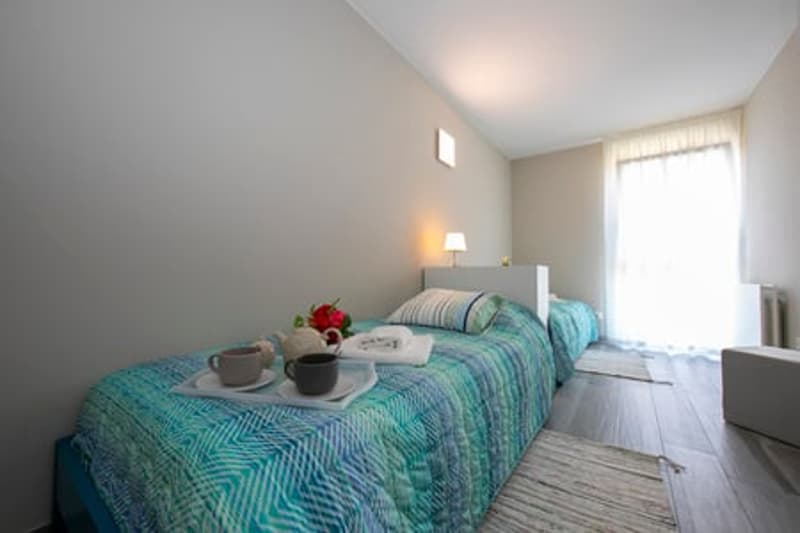 bedroom with two beds, table with teapots, curtains, lamps, small flower vase, blankets