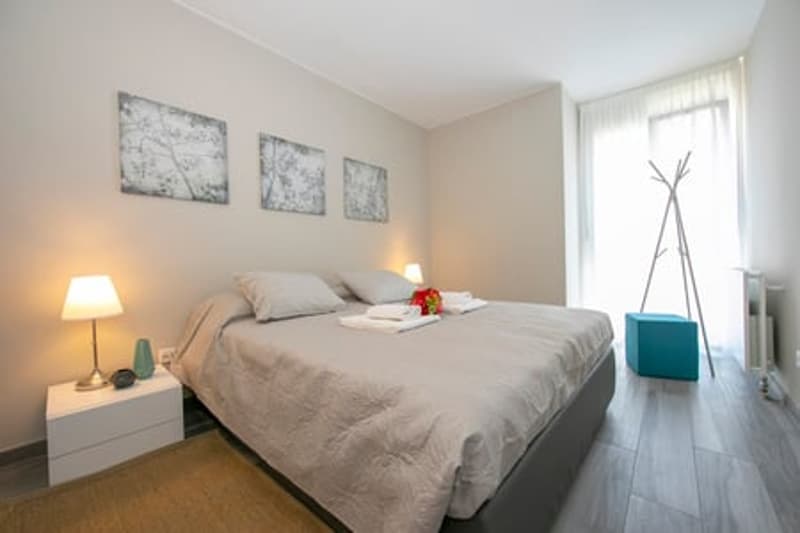 A bedroom with white walls, wooden floor, a bed with gray bedding, two nightstands with lamps, a rug, a coat rack, and a window with white curtains
