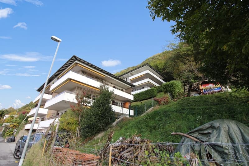 Multi-story building, white exterior, balconies, green surroundings, parking area