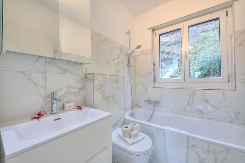 Modern bathroom with marble tiles, white fixtures, separate shower area, bathtub, toilet, vanity with sink, and natural light from window.