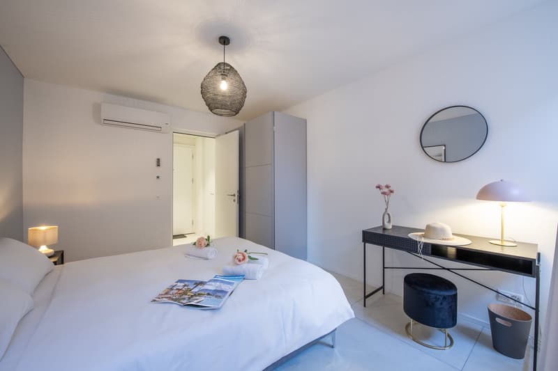 White bed with white sheets, 2 bedside lamps, air conditioner, round mirror, desk with hat and lamp, small table with trash can, cabinet, flower vase, flowers on bed, white tile flooring