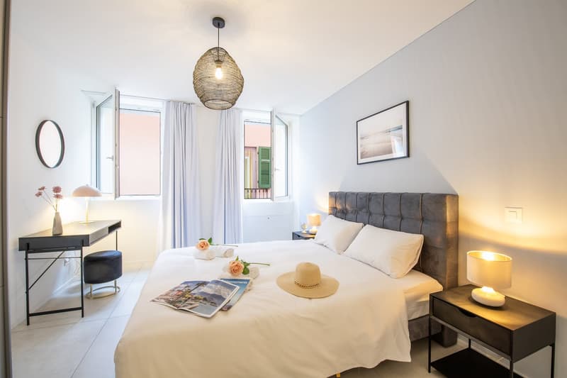King size bed, white walls, two nightstands with lamps, desk with round mirror, round hanging lamp, window with balcony, picture frame