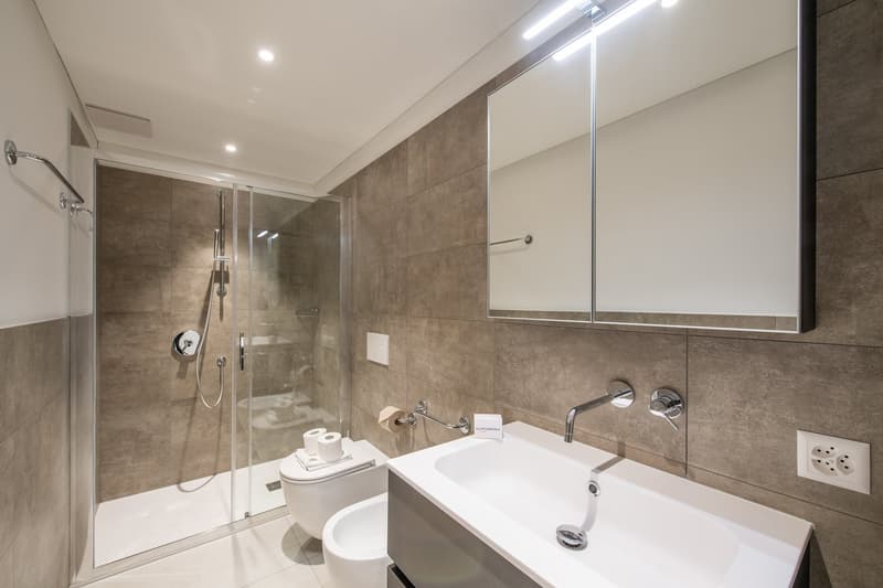 Modern tiled bathroom with glass shower, white sink, mirror, toilet, and bidet.