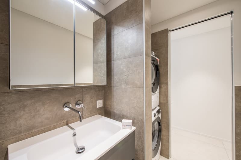 Stylish bathroom with large mirror, gray tiles, white sink, faucet, and built-in washing machine in adjacent room