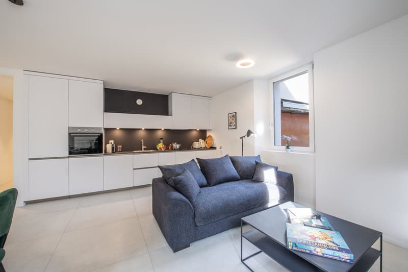 Modern kitchen, white cabinets, black countertop, built-in oven, sink, countertop with books and board game, couch, floor lamp, window with view of outside