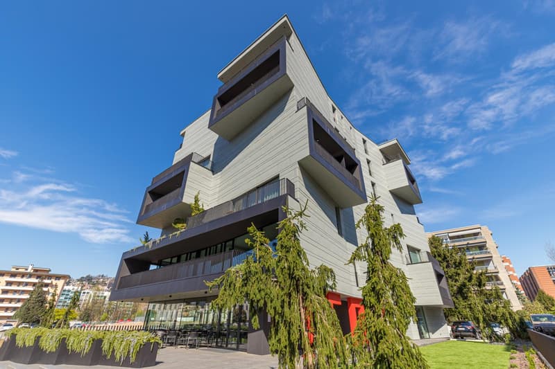 Modern, angular building with multiple balconies, glass facade, green plants, and trees in the foreground
