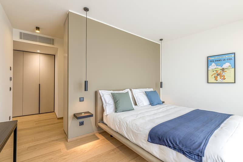 Modern bedroom with wooden floor, white bed, decorative pillows, mounted light fixtures, wooden table, and picture frame on the wall